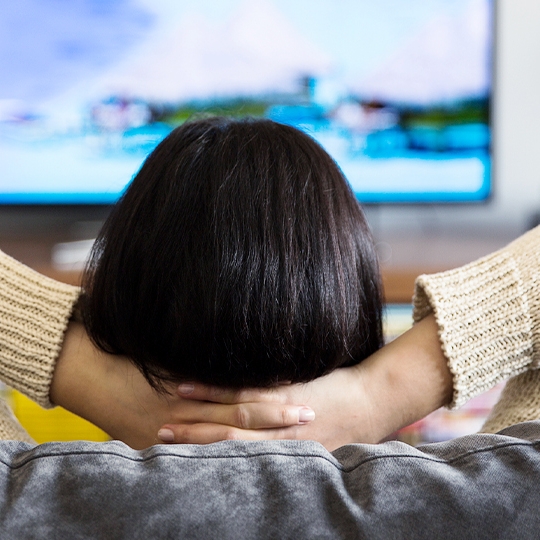 Frau von hinten mit verschränkten Armen schaut entspannt Fernsehen.