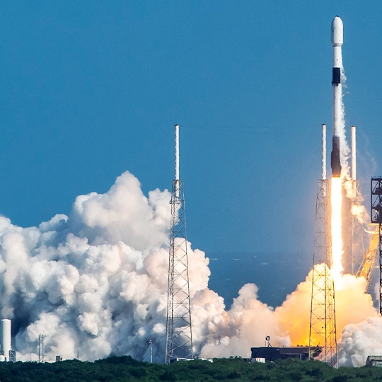 Eine Rakete hebt von einer Startrampe ab, umgeben von großen weißen Rauchwolken. Der Himmel ist klar und blau. Das Bild vermittelt Dynamik und Fortschritt.