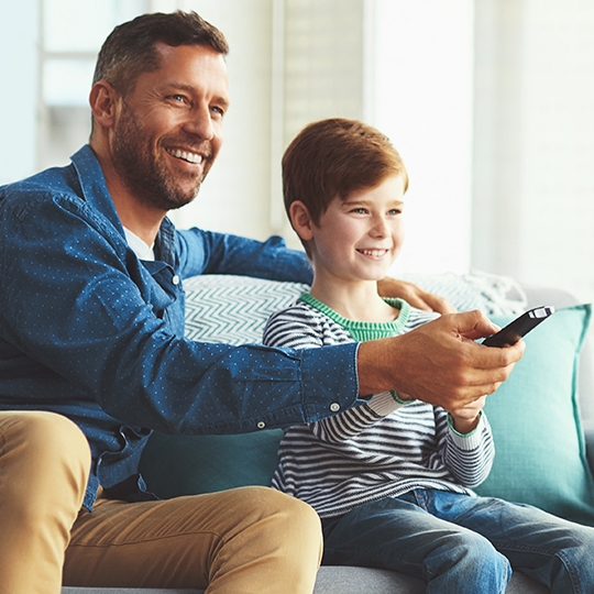 Ein Mann und ein Junge sitzen lächelnd auf einem Sofa in einem hellen Wohnzimmer und schauen gemeinsam fern. Der Mann hält eine Fernbedienung. Das Bild vermittelt eine familiäre und gemütliche Atmosphäre.