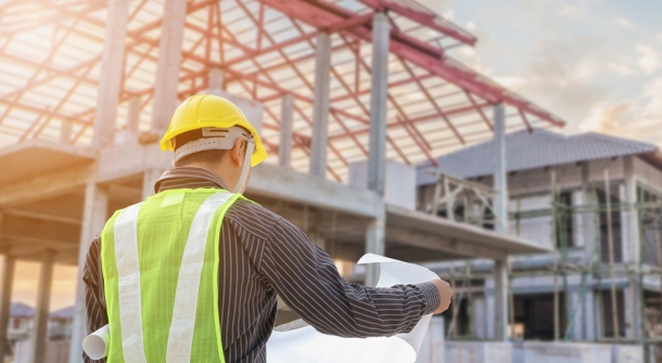 Ein Bauingenieur oder Bauleiter in Schutzkleidung, inklusive gelbem Helm und leuchtender Warnweste, betrachtet Baupläne vor einer Baustelle. Im Hintergrund ist ein unvollendetes Gebäude mit Gerüsten und einem Dach im Bau zu sehen. Die Szene spielt bei Sonnenuntergang, was eine warme und angenehme Lichtstimmung erzeugt
